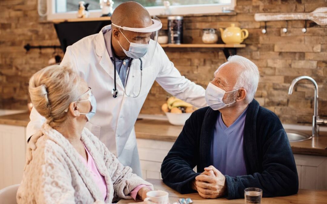 A doctor wearing a mask and face shield leans over a kitchen table, speaking warmly to an elderly couple also in masks. The room is cozy with warm lighting.