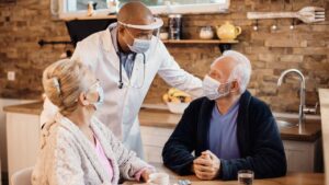 A doctor wearing a mask and face shield leans over a kitchen table, speaking warmly to an elderly couple also in masks. The room is cozy with warm lighting.