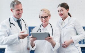 Three doctors in white coats and stethoscopes are smiling and looking at a tablet. They appear engaged and happy in a clinical setting.