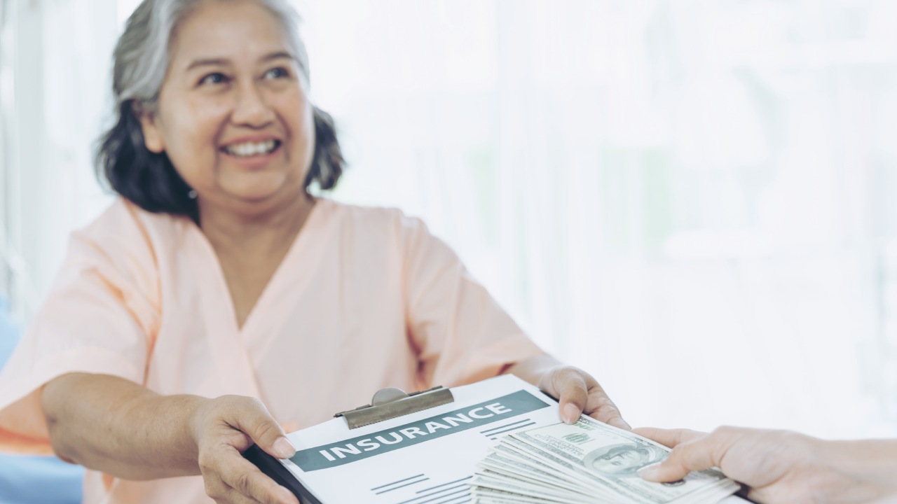 Smiling elderly woman in a light pink blouse hands over an insurance document on a clipboard while another hand offers a stack of dollar bills.