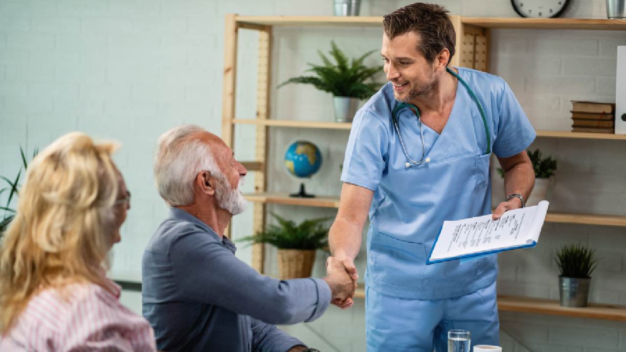A smiling healthcare professional in blue scrubs shakes hands with an elderly man, conveying warmth. A woman sits nearby in a cozy, plant-filled room.